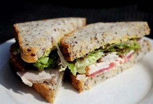 A sandwich made using seeded sliced bread, filled with chicken, tomato and lettuce, cut diagonally into two halves and placed on a white plate