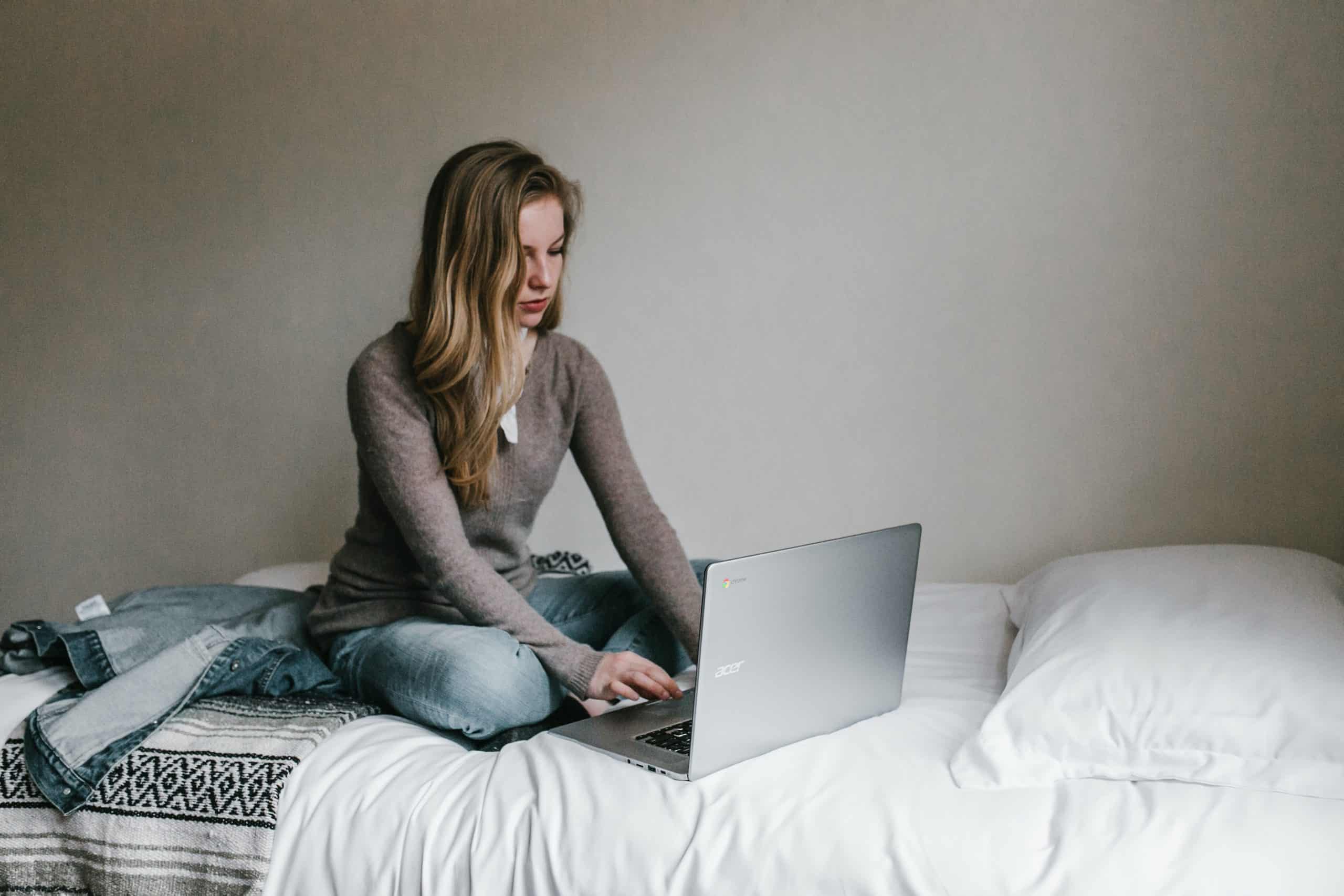 blonde female sitting on a bed typing on a silver laptop