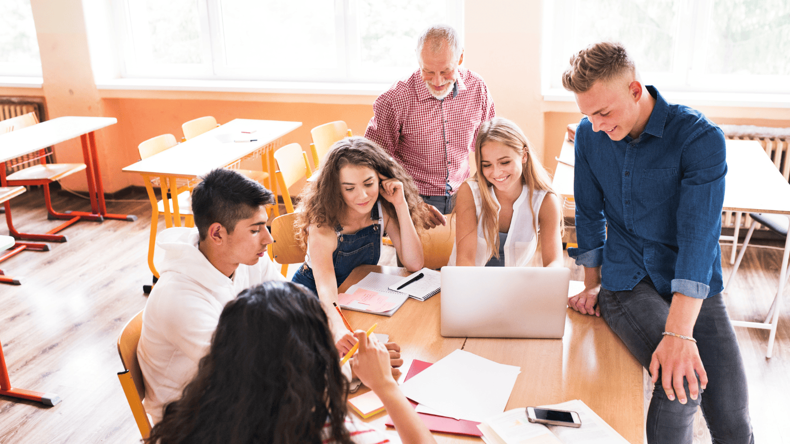 Teacher and teenage students gathered around a desk in the classroom and looking at a laptop and papers