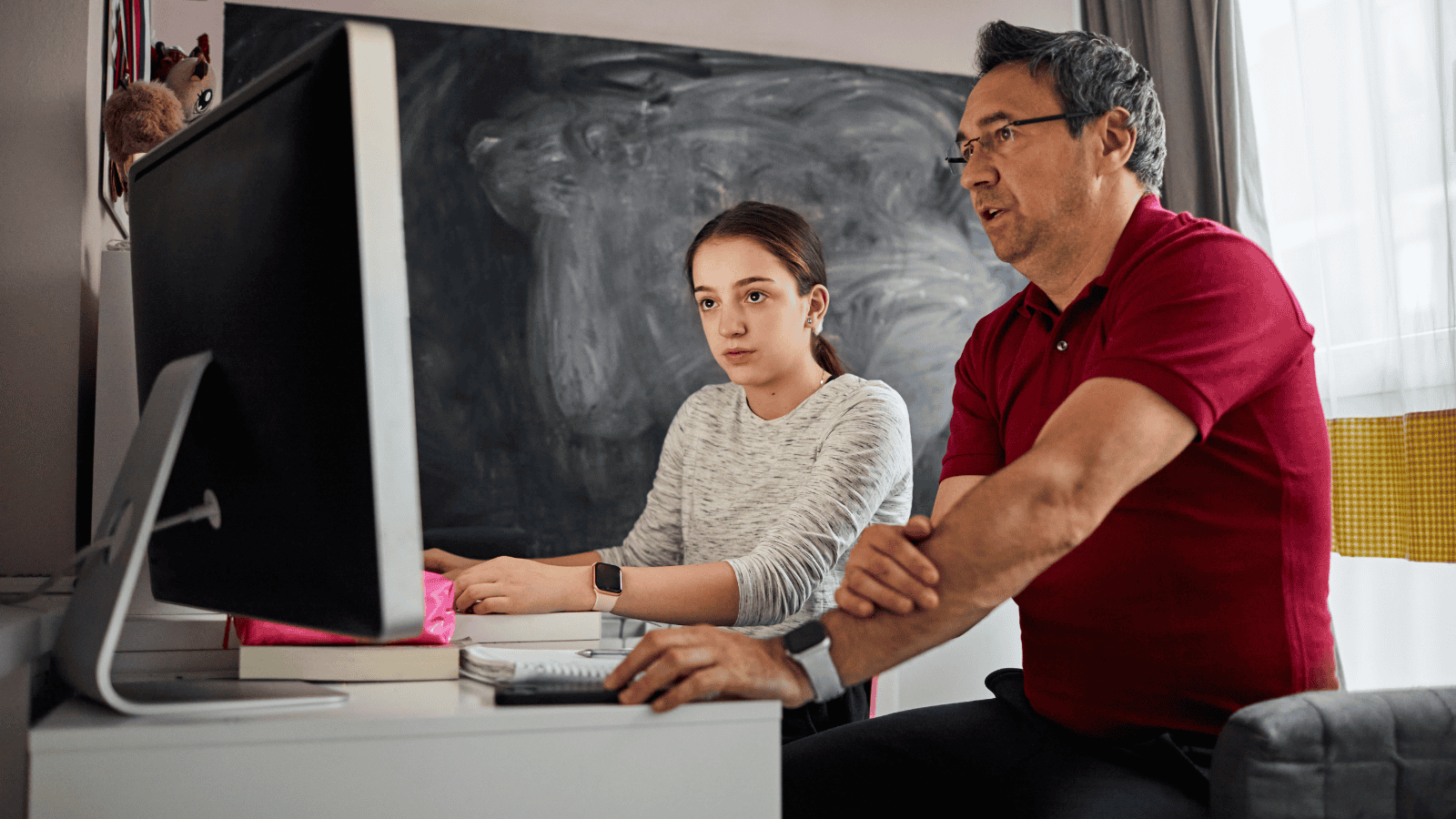 Father and daughter sitting together at a desk looking at a computer screen and with notebook on the table.